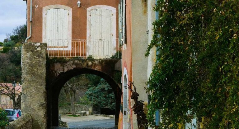 Porche voûté en pierre sous une maison aux volets clairs avec une sculpture en métal dans un village provençal